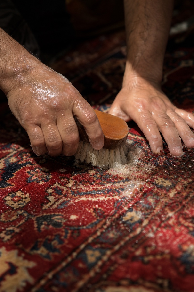 Hands working a wash brush through the wet pile of a Persian rug